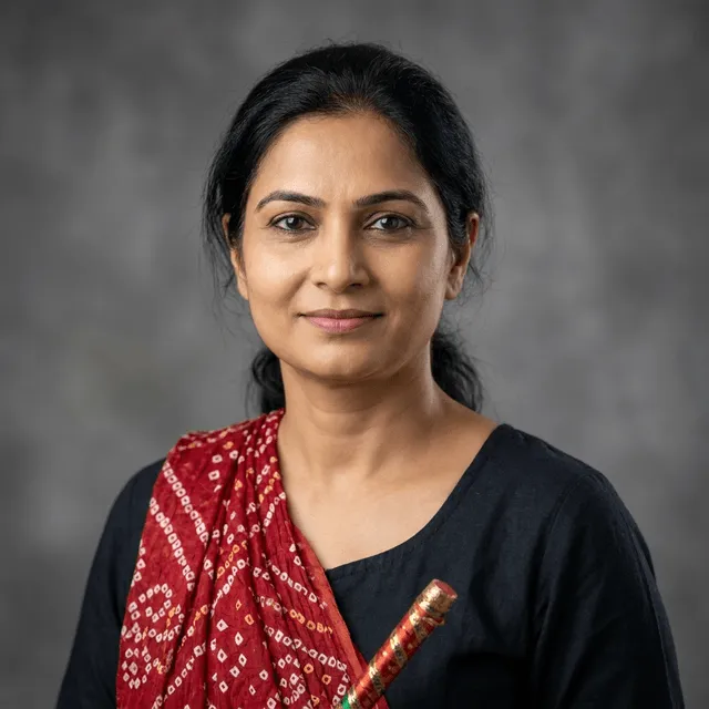 Middle-aged woman with dark hair pulled back, wearing a black top with a red and white patterned scarf draped over one shoulder and holding a decorated stick, with a slight smile.