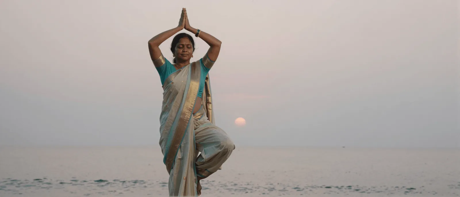 A woman in a saree holding a yoga tree pose on the beach at sunrise