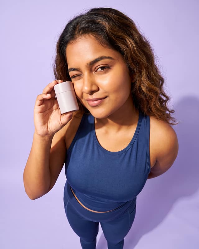Skincare launch: Serum bottle held to the cheek, lilac seamless, a wink.